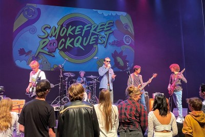 The band, High Voltage perform on stage at Rockquest, with three people on guitar, one on drums and one singing as they hold a microphone.