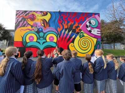 Photo is taken from behind Year 8 students as stand in forecourt area outside the Christchurch Art Gallery looking at the colourful mural Kuīni of the Worlds which was painted by Xoë Hall.