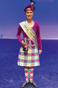 Millie stands on stage in her Highland Dancing outfit, with a sash around her body and holding a trophy.