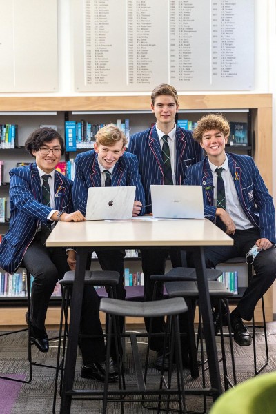 Luke Zhu, Tom Edwards, Corin Simcock and Toby Harvie sit behind a table with two laptops in the College library and smile for a photo.