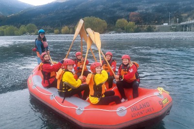 A group of students are in a raft in a body of water, with lifejackets and helmets on, their paddles in the air, as they smile for a photo.