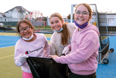 Three girls from the Preparatory School stand on the turf with a rubbish bag open and smile for a photo.