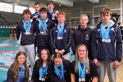 The St Andrew's College swimming team bunch up for a group photo in front of the pool. They wear their St Andrew's College tracksuits and have the medal they have won around their necks.